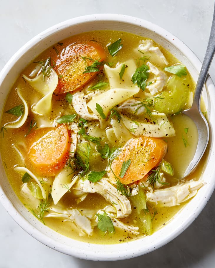 Close-up overhead shot of turkey noodle soup in a white bowl garnished with fresh parsley and cracked black pepper, with a linen napkin and crusty bread on the side